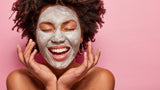 Cheerful young woman has Afro hairstyle, touches cheeks, has clay mask on face,